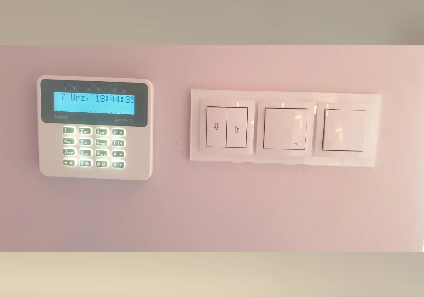 Bezprzewodowy manipulator Satel PRF-LCD systemu Perfecta zamontowany w linii z włącznikami światła na ozdobnej różowej ścianie – estetyka Winkam Rybnik.